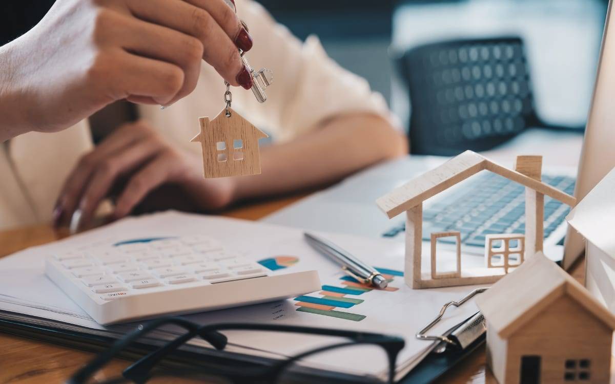 A hand holding house-shaped keys over wooden model houses, a calculator and property charts
