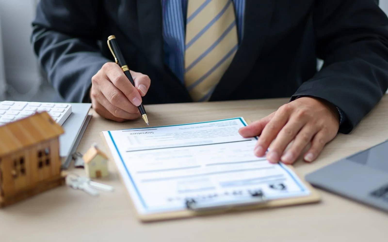 A man in a suit signing an application form on a clipboard, with a set of keys and a small wooden house model on the desk
