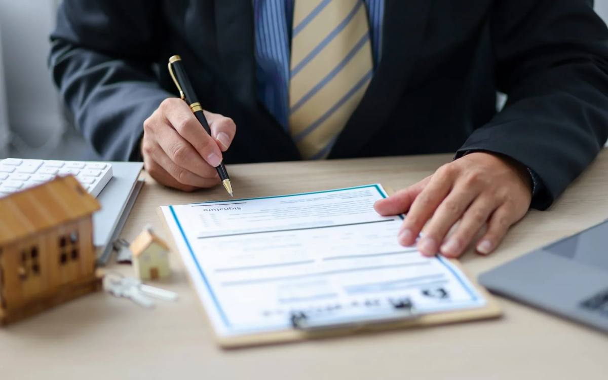 A man in a suit signing an application form on a clipboard, with a set of keys and a small wooden house model on the desk